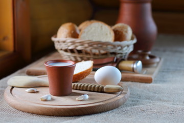 A cup of milk  and a wicker basket with bread. A clay cup with milk and a wicker basket with bread are on the table near the window in the village house.