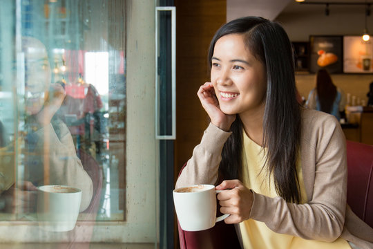 Asian woman sitting next to glass window inside coffee shop with cup of hot coffee in left hand