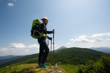 A young woman in black with green backpack standing on top of a hill enjoying an amazing view of the mountains.