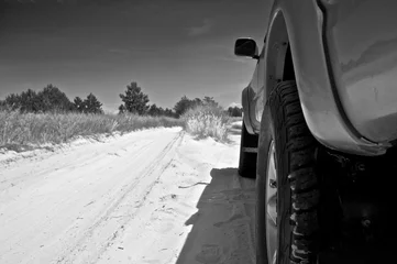  Picture taken from behind the rear wheel of a pickup truck parked on the side of a dirt road. Grass and trees seen in background. Black and white.  © Oleksii Klonkin