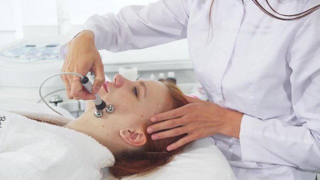 Cosmetologist using iontophoresis for client's face. Female hands moving ball head of machine along girl's chin. Pretty caucasian woman getting galvanic facial treatment