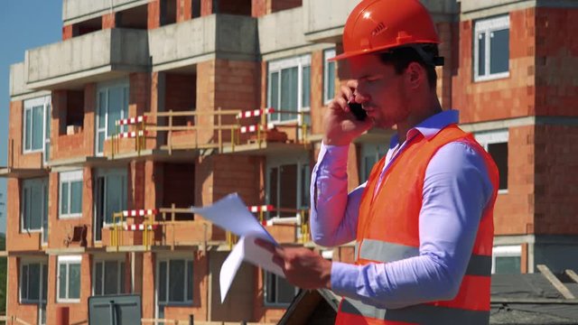 Construction worker phones with the smartphone and holds some papers in front of building site