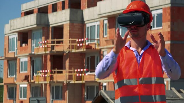 Construction worker uses virtual reality glasses and plans something with hands in front of building site 