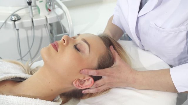 Cosmetologist dressed in white coat massaging client's head. Female beautician touching woman's ears and temples. Pretty blond girl getting accupressure treatment at the beauty salon