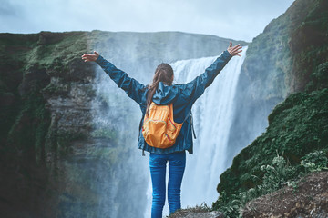 Girl in waterproof clothing stands on the cliff on background of Skogafoss waterfall in Iceland. back view, woman holds hands up