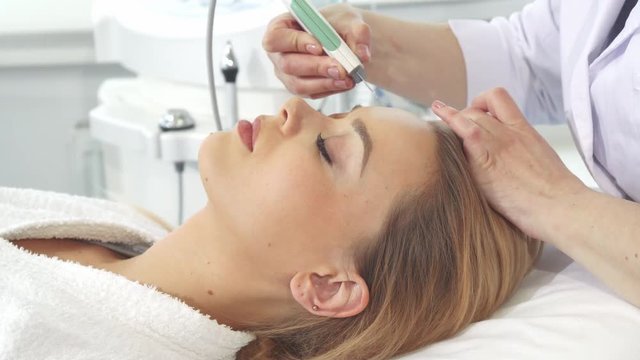 Attractive female client getting ultrasonic peelling for her face. Professional beautician moving scrubber along girl's forehead. Cosmetologist performing deep cleaning of woman's facial skin