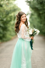 Fototapeta na wymiar Bride in beautiful wedding dress stands on forest road, smiles and looks back.
