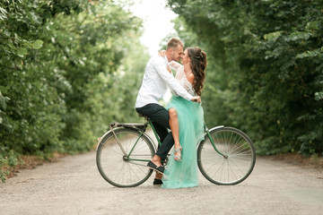 Bride and groom sit on bicycle on forest road, embrace, laugh and kiss.