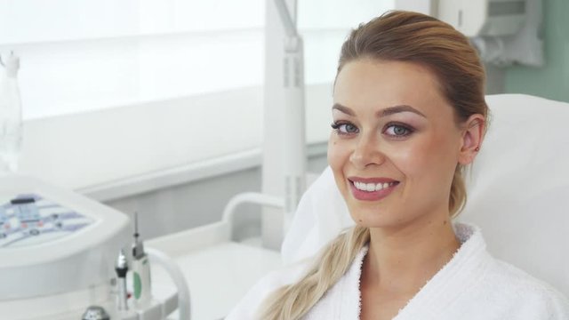Pretty blond woman smiling for the camera at the beauty salon. Attractive young woman sitting on the chair against background of some cosmetology equipment. Female caucasian client showing the result