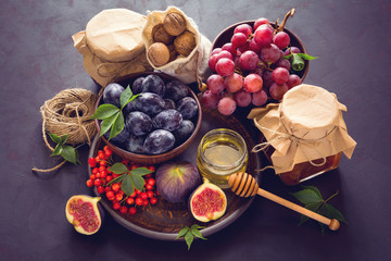 Harvest set:  plums, grapes, figs, viburnum berry, walnuts, honey and jam glasses on dark wooden desk. Still life. Toned image