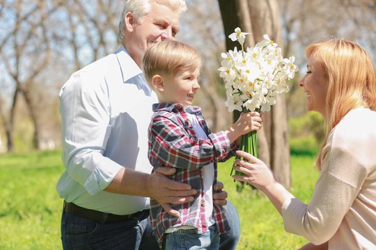 Cute little boy with grandfather giving bouquet of flowers to his grandmother in spring park on sunny day