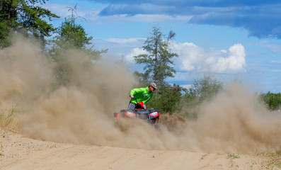 A 16 year old teenaged boy spinning circles creating a cloud of dust on a quad motorbike on a sandy road lined by trees in a summer countryside landscape