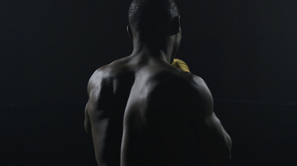 Rear view of muscular man boxing on black background. Afro american young male boxer practicing shadow boxing. Boxer in yellow Boxing gloves - obrazy, fototapety, plakaty