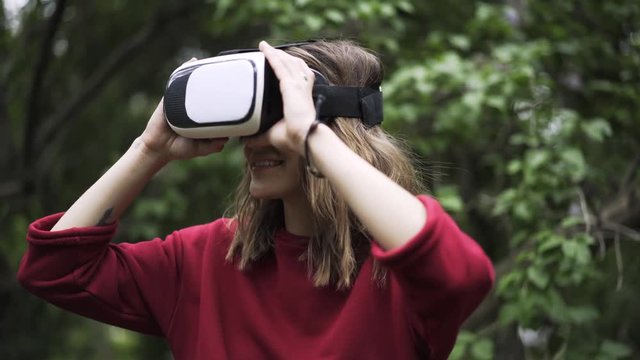 Young woman wearing a red sweater is using VR glasses while being at a park near large tree. Handheld real time close up shot