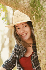 Beautiful japanese descendant woman on a nature scene. Portrait of a smiling woman wearing hat behind a tree trunk.