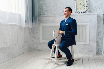 Fashionable cheerful man in a blue suit is sitting on a toy horse. Adult acting like a child concept.