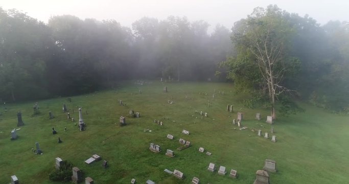 A forward high angle aerial above a historic cemetery in the foggy woods.  	
