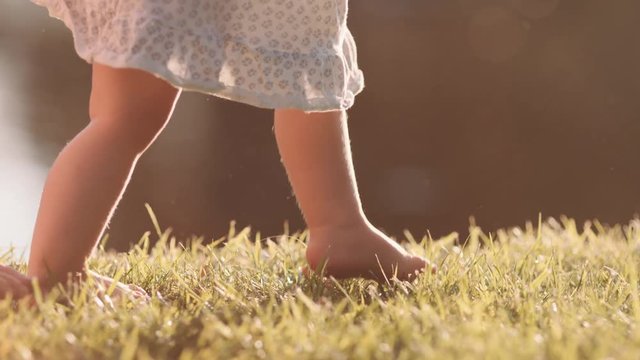 Little baby learns to walk. First Steps. Slow Motion 120 fps, 4K. Barefoot child does the first steps on a green grass in summer. Close up on feet. Happy childhood and Maternity. Family