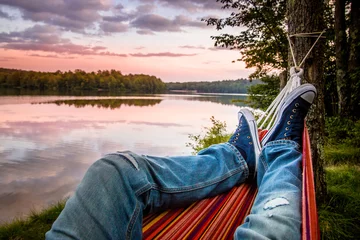 Tuinposter Zomerkamperen aan het meer. Jonge man met jeans en sneakers ontspannen in de hangmat bij zonsondergang. © Nicolae Merceanu
