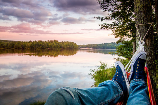 Summer camping by the lake. Young man wearing jeans and sneakers relaxing in the hammock at sunset. 