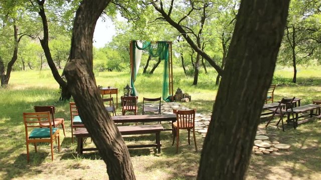 Wedding arch in rustic style with a green and white cloth with old chairs and benches in the woods.