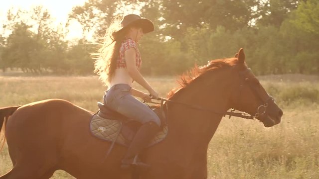 Two young girls with long hair and a cowboy hat galloping on horseback in the field at sunset, slow motion. Two girls riding a horse in a field at sunset.