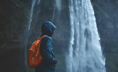 Girl in waterproof clothing stands under the Seljalandsfoss waterfall in Iceland. back view, woman with small orange backpack