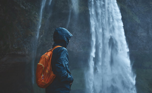 Girl in waterproof clothing stands under the Seljalandsfoss waterfall in Iceland. back view, woman with small orange backpack