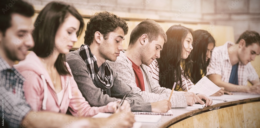 Poster students writing notes while sitting in row at lecture hall