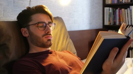 Young handsome man lying in bed and reading a book in his bedroom at home at night - Powered by Adobe