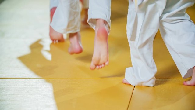 Kids are jumping up and then they are kneeling down during a warm up judo practise. All of them are wearing white uniforms.