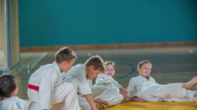 Kids are lying down on the ground in a school gym when they are warming up for a judo practise. They are wearing white unifoms.
