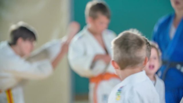Children are lifting their legs up when they are warming up before judo practise in class. They are wearing white judo uniforms.