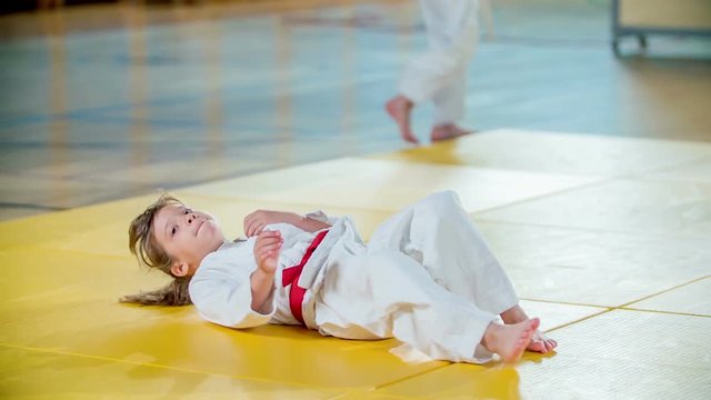 A young girl is performing one of the moves in judo. All the kids are having practise after school. They are dressed in white uniforms and are wearing belts of different colours.