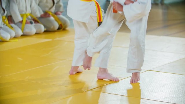 Two students are practising two of the judo moves on a yellow mat in front of their friends. All of them are wearing white judo uniforms.