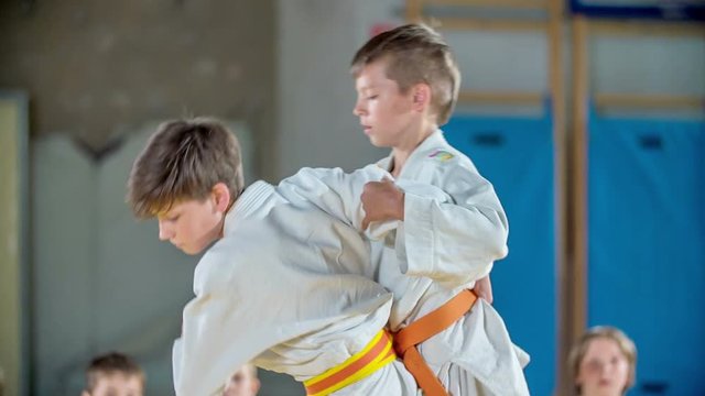 One boy throws the other boy over his shoulder during judo practise. They are wearing white uniforms and different colour of their belts.