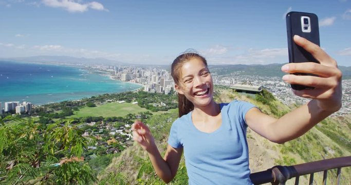 Cheerful female hiker taking selfie through smartphone against Waikiki Beach and Honolulu. Woman is enjoying vacation at Diamond Head State Monument. She is gesturing peace sign while photographing.