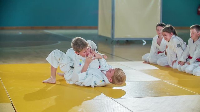 Two boys are fighting in judo on a yellow mat in the school gym. The rest of the students are watching them.