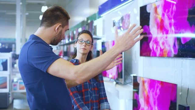In the Electronics Store Professional Consultant Provides Expert Advice to a Young Woman Who Looks for a  New 4K UHD TV to Buy. 