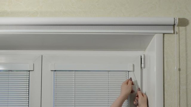 Female hands with beautiful manicure lower horizontal blinds at a window with a frame from white plastic