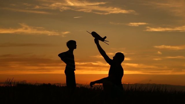 A happy family. The boy on his father's shoulders is playing with an airplane toy. Silhouette at sunset by the lake. at this time a real airplane is flying in the sky