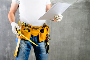 unidentified builder standing in white gloves with a tool belt with construction tools and holding roulette and the project plane against grey background. DIY tools and manual work concept