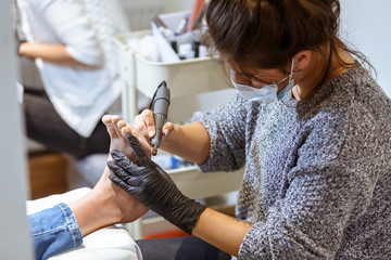 A young white manicurist girl makes a pedicure electric nail drill machine to the client in the workplace. On her face a protective dressing, on her hands protective gloves. Close view.