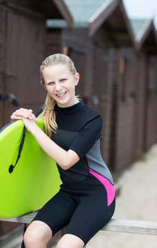 Portrait Of Girl By Beach Huts In Wetsuit Holding Bodyboard