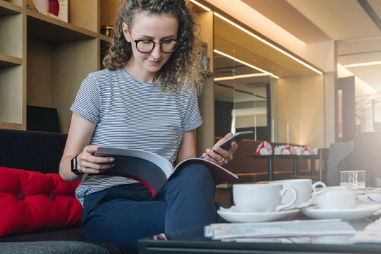 Young businesswoman sits on sofa in hotel lobby, restaurant, cafe and reads book, magazine, catalog.