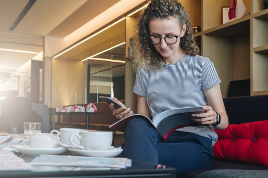 Young businesswoman sits on sofa in hotel lobby, restaurant, cafe, drinks coffee and reads book, magazine, catalog.