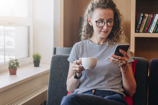 Young businesswoman sits on sofa,uses smartphone,working, drinking coffee.Hipster girl is chatting,blogging,checking email