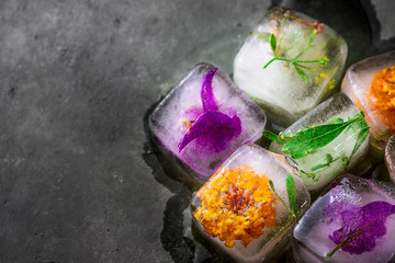 Ice cubes with frozen colorful flowers herbs and plants melting on dark stone background. Beauty, face skin care concept. Rejuvenation, ayurveda. Wellness spa.