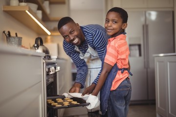 Father taking tray of fresh cookies out of oven with son in