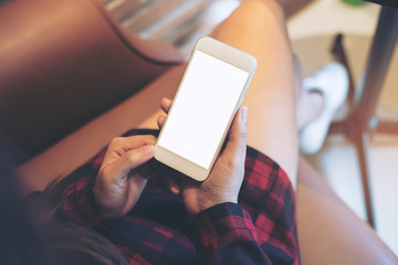 Mockup top view image of woman sitting cross legged and holding white mobile phone with blank screen on thigh in modern cafe